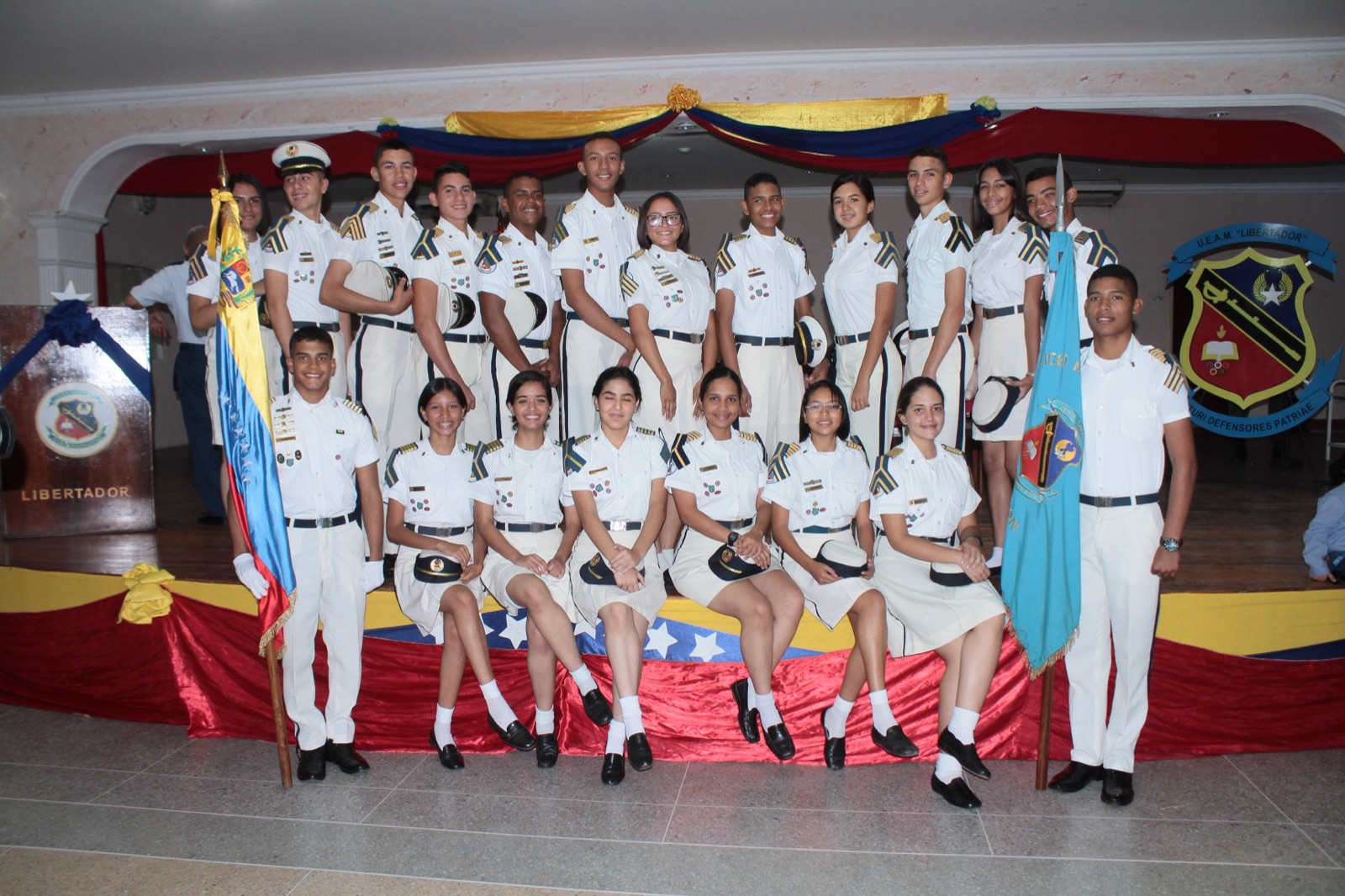 Cadetes en uniforme de gala posando frente a las banderas y escudo institucional